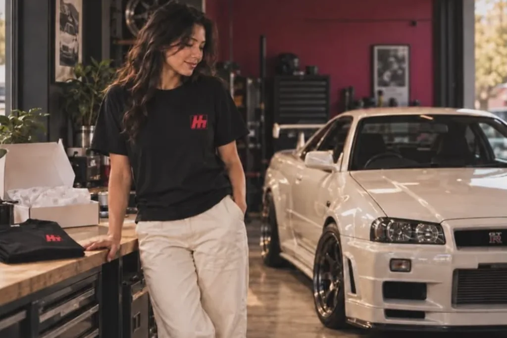 A woman in a black ClutchCloth T shirt leaning on a garage workbench beside a white sports car